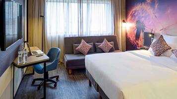 Habitación de hotel con cama, escritorio con una computadora portátil, silla, sofá y un ventanal con cortinas.