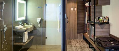 A modern hotel room with a glass shower enclosure, a separate bathroom with a sink and mirror, a bed with a white comforter, and a wooden vanity with a mirror and storage.