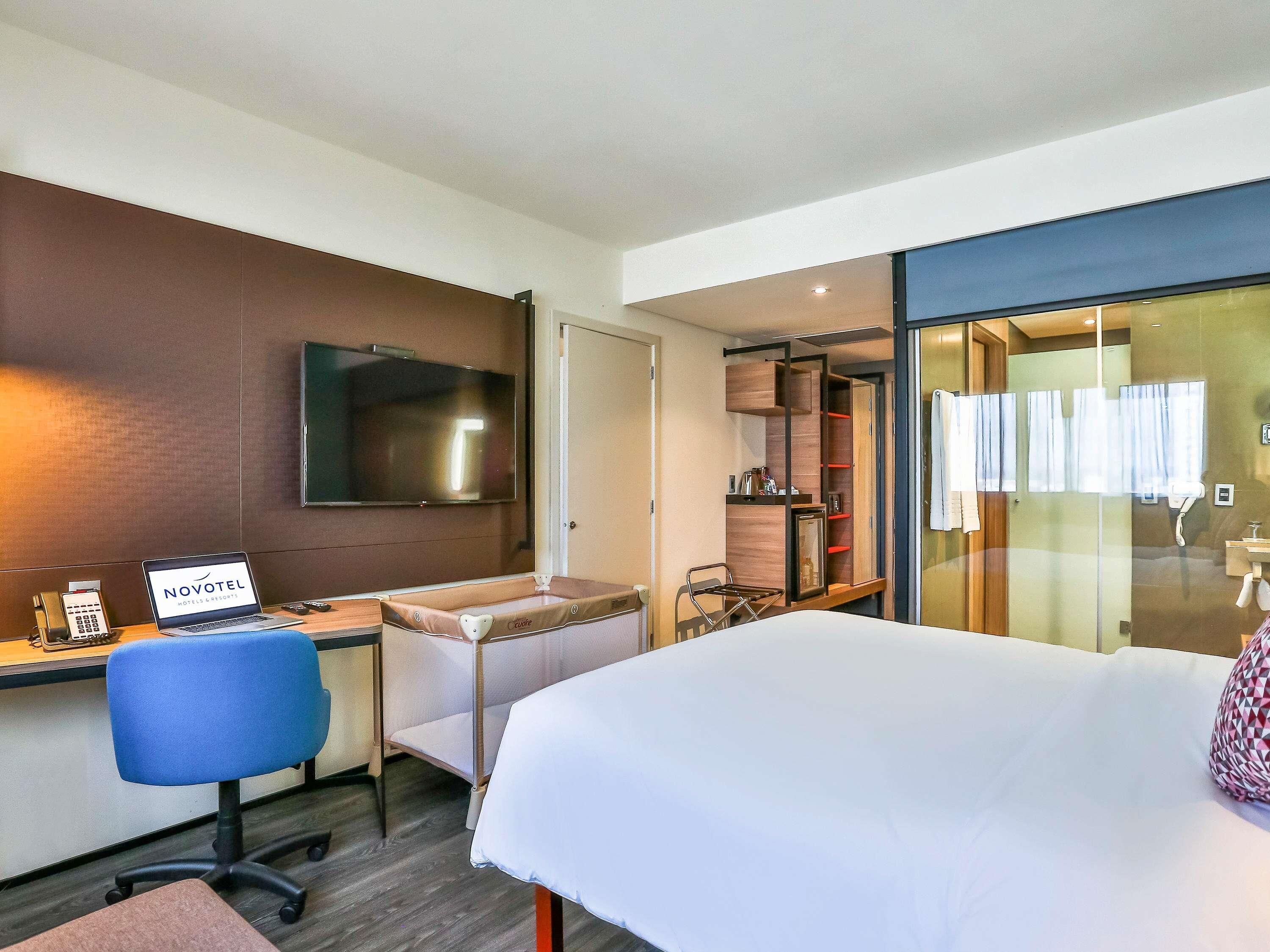 A hotel room with a bed, a desk with a laptop, a chair, a TV, and a sliding door leading to a bathroom.