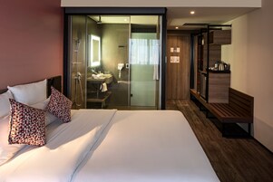 Una habitación de hotel moderna con una cama grande, ducha con cerramiento de vidrio, un banco de madera y una pequeña cocina.