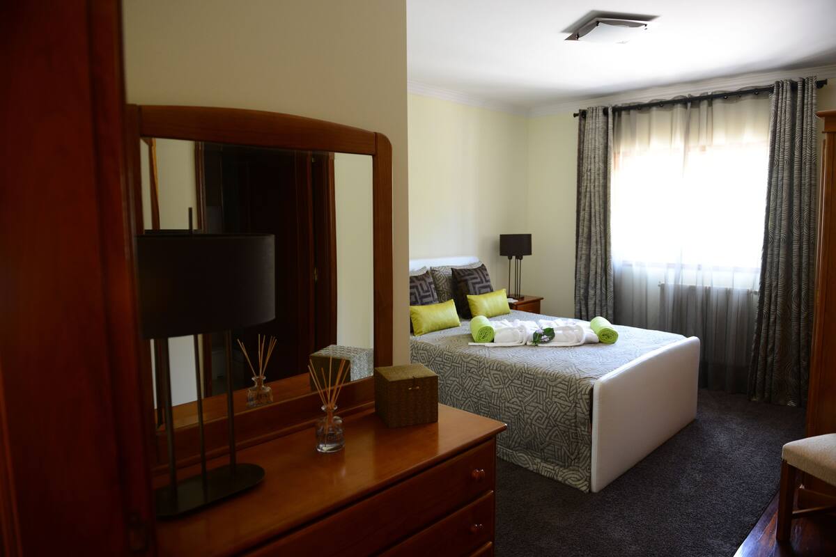 A hotel room with a large bed, a dresser with a mirror, a bedside table with a lamp, and a window with curtains.