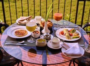 A round glass table with a variety of food and drinks, including a plate of pasta, a bowl of soup, and a glass of juice, set outdoors with a grassy background. - Sterling Kanha (Bichhiya)