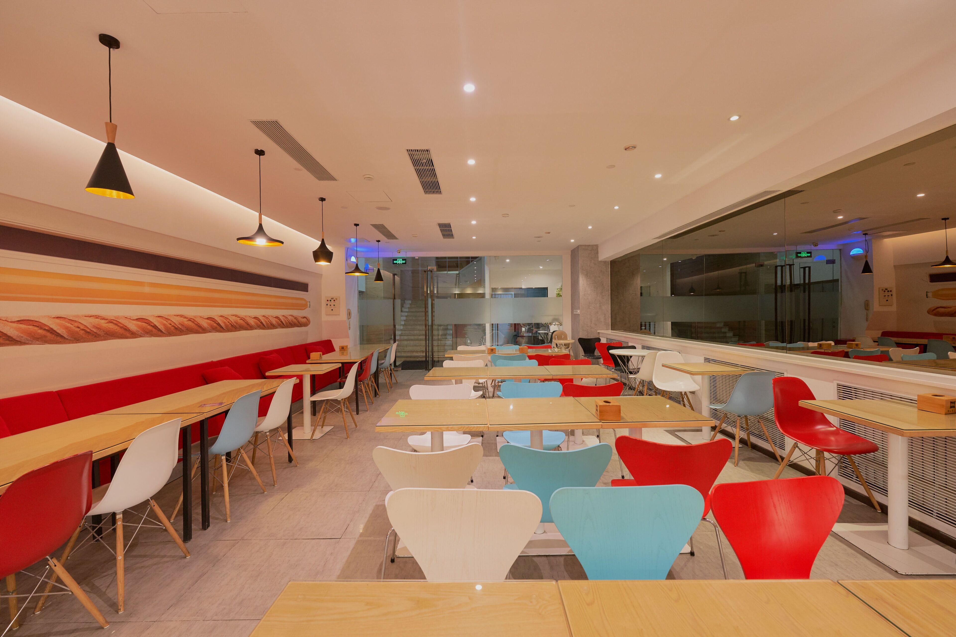A modern restaurant with long wooden tables and colorful chairs, a bright ceiling with pendant lights, and a large window on one side.
