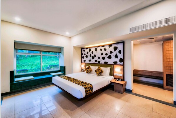 Una habitación de hotel moderna con una cama grande, un ventanal con vistas a zonas verdes, un banco integrado y un cabecero con un diseño único.