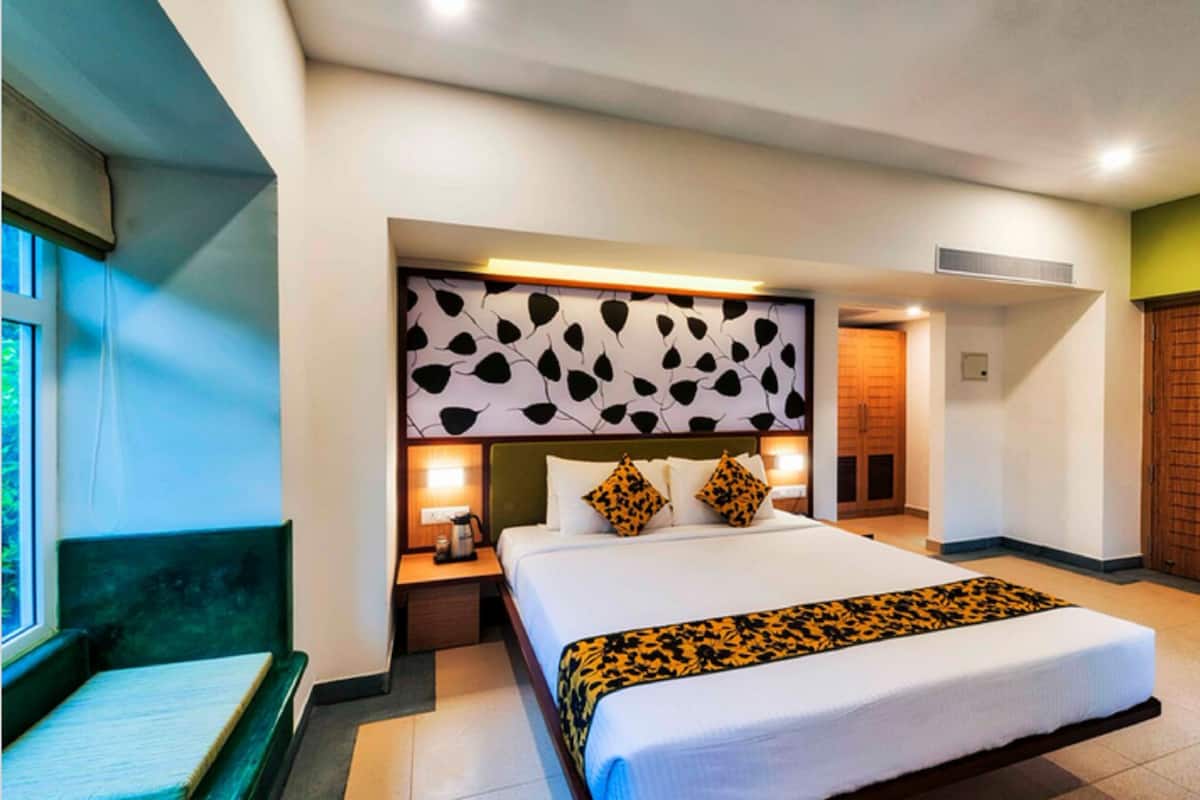 A hotel room with a large bed, a bedside table with a lamp, a window with a view, and a green bench by the window.