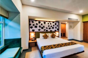 A hotel room with a large bed, a bedside table with a lamp, a window with a view, and a green bench by the window. - Sterling Wayanad (Sulthan Bathery)