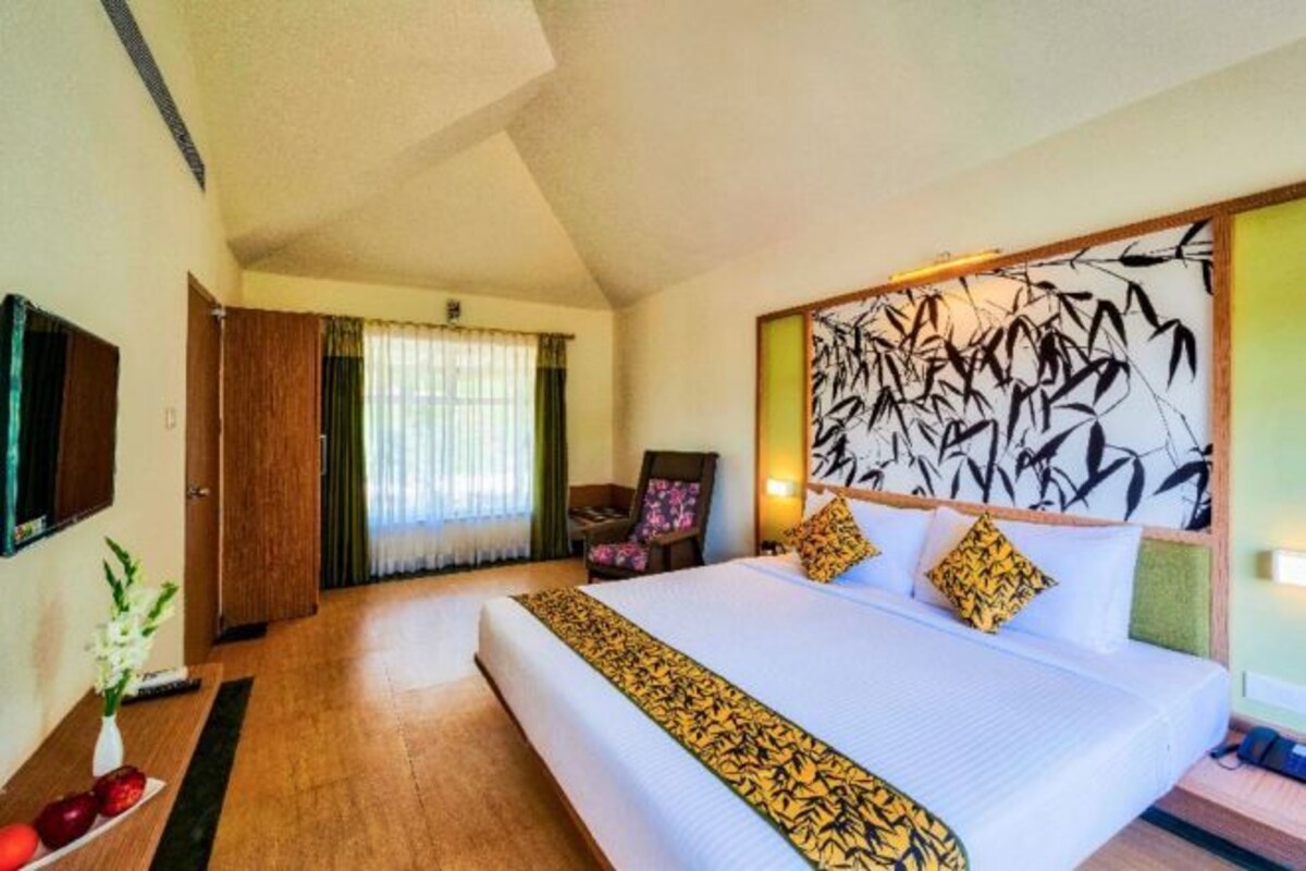 Habitación de hotel con una cama grande, un televisor montado en la pared, un escritorio con un florero y una ventana con cortinas.