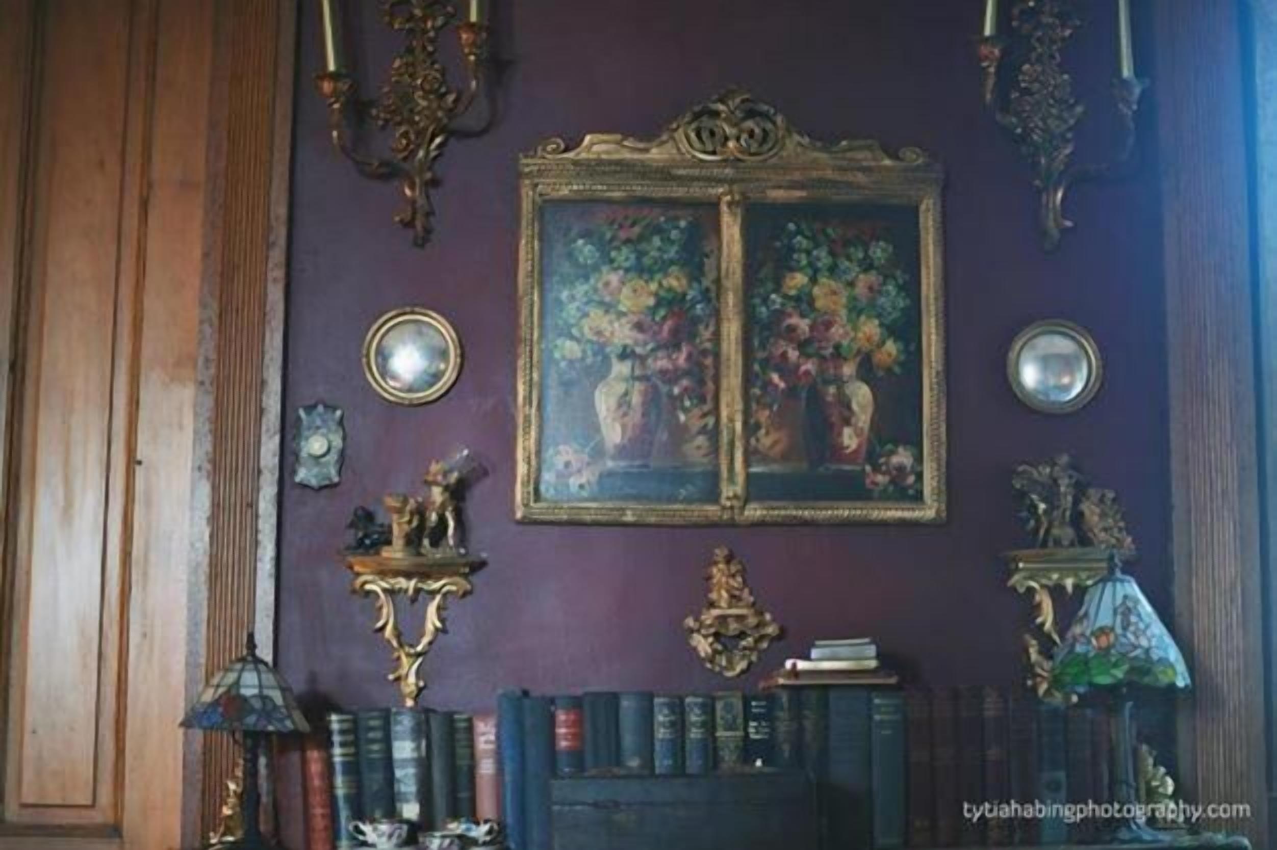A room with a purple wall, a framed floral painting, a bookshelf with books, and ornate wall sconces.