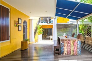 A covered outdoor area with a wooden deck, a colorful table, and a staircase leading to an upper level. - Bario Hotel (Willemstad)