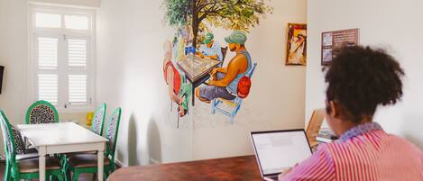 Un comedor con un mural de gente alrededor de una mesa, una mesa blanca, sillas verdes y un ordenador portátil sobre la mesa.