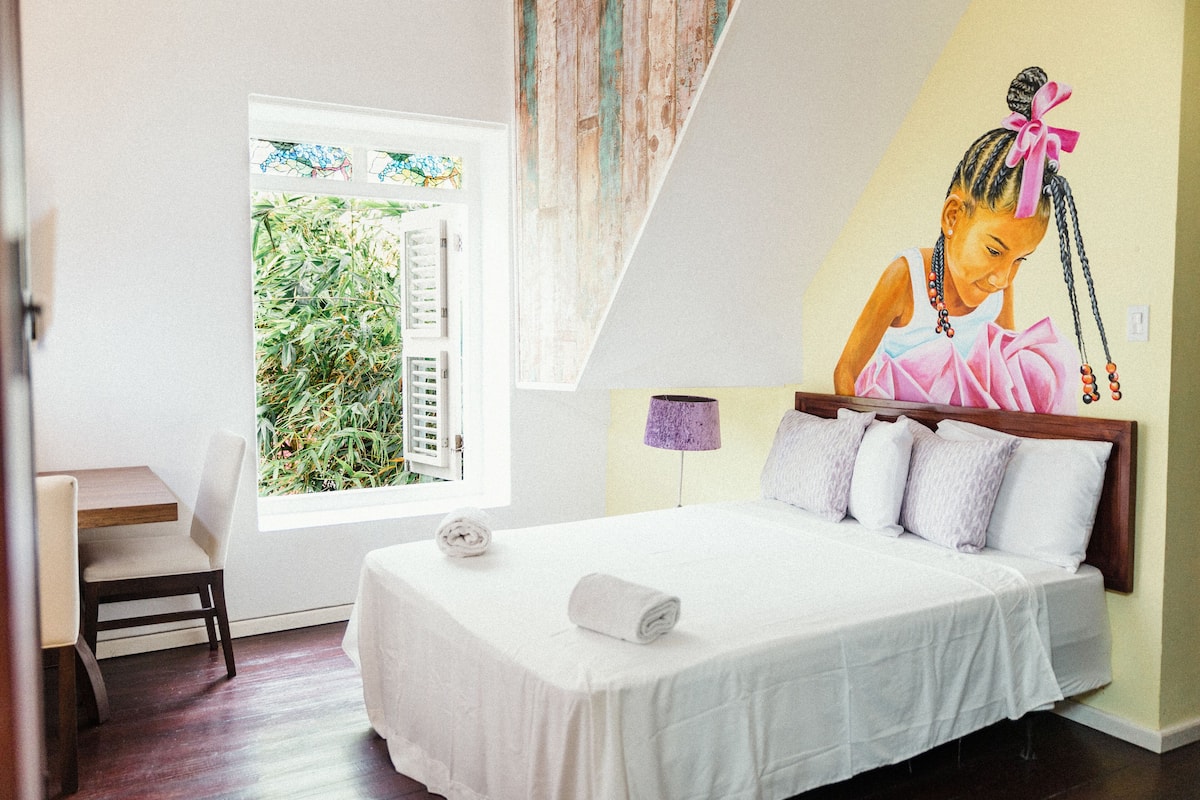 A bedroom with a bed, a chair, a window with a view of greenery, and a wall with a mural.