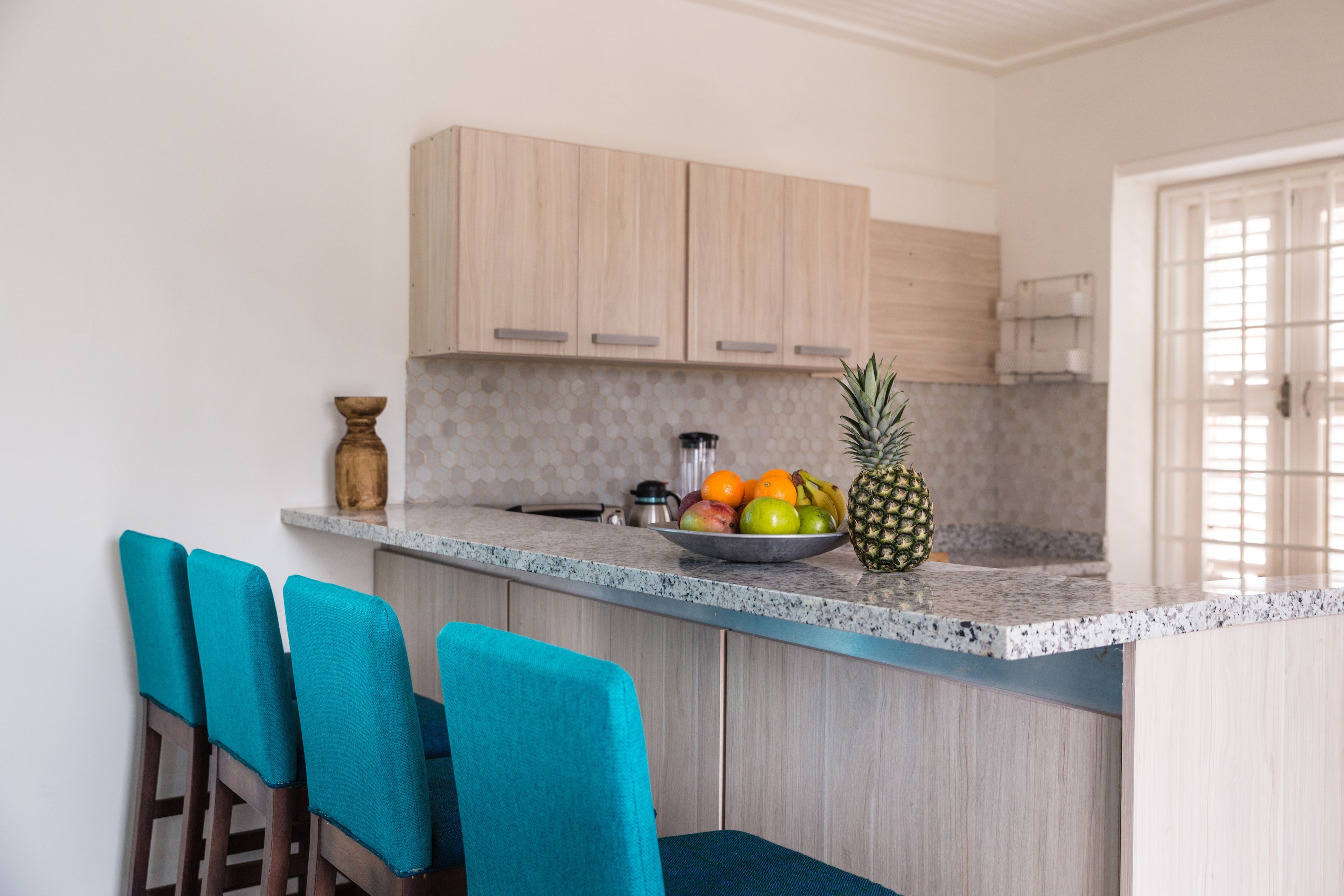 A kitchen with light wood cabinets, a granite countertop, and a fruit bowl containing apples, oranges, and a pineapple. There are teal chairs and a small vase on the counter.