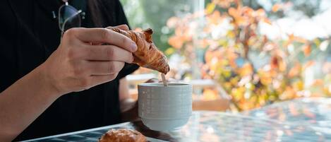 A person holding a croissant, a cup, and a napkin on a table with a window view.