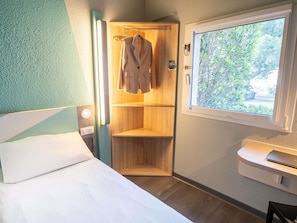 A bedroom with a bed, a wooden wardrobe, a window with a view of greenery, and a bedside table.
