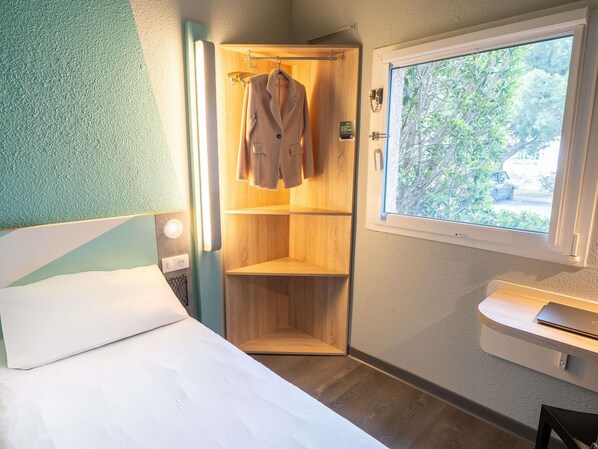 A bedroom with a bed, a wooden wardrobe, a window with a view of greenery, and a bedside table. - ibis budget Narbonne Est (Narbonne)