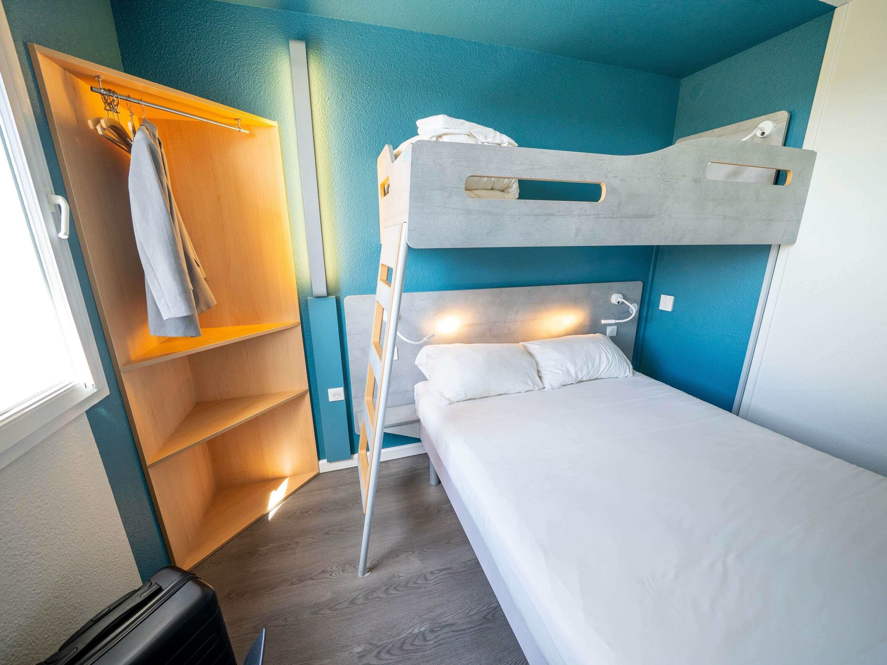 Uma cama beliche com encosto, um guarda-roupa de madeira com cabides e uma mala no chão.