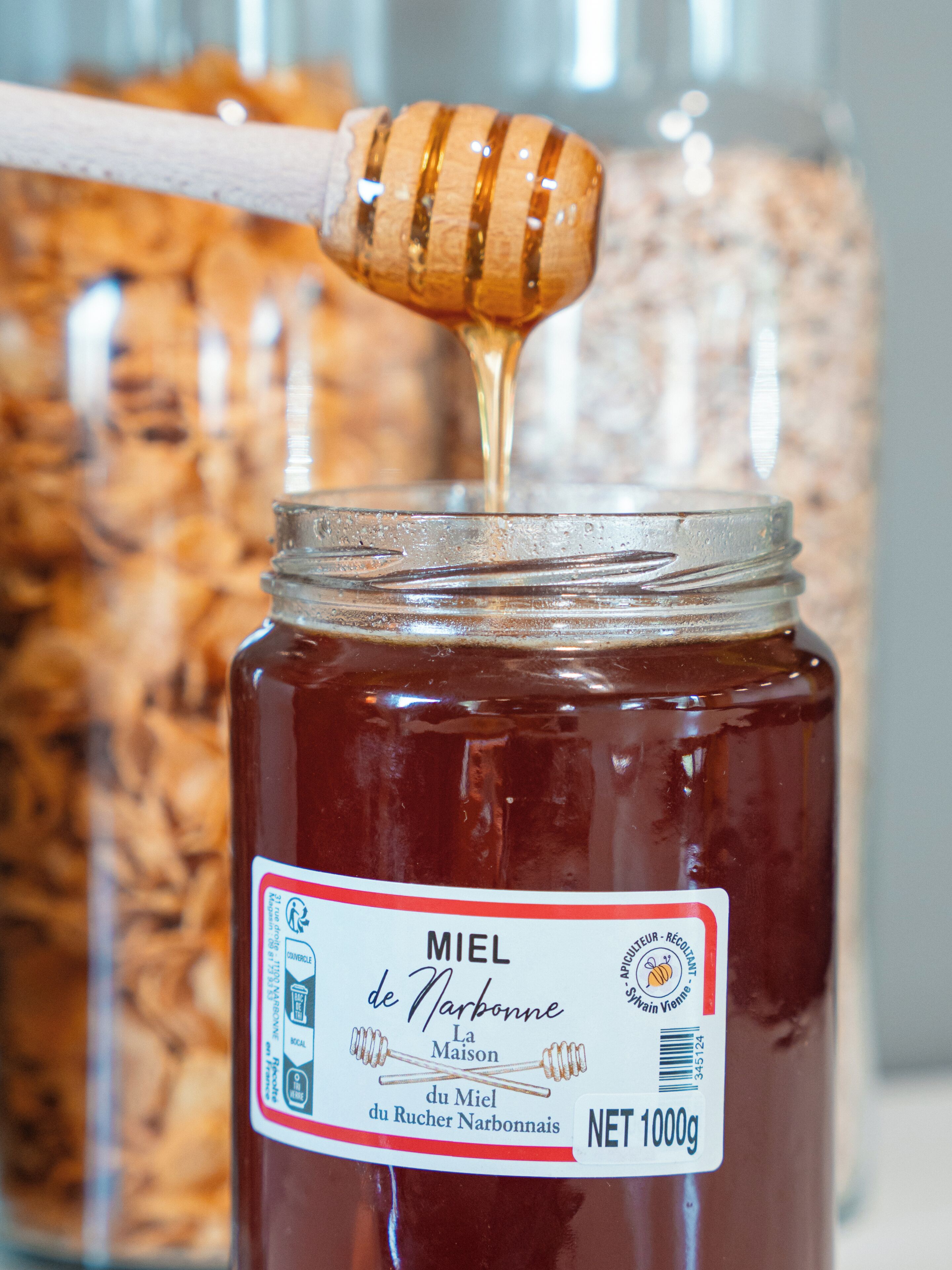 Un pot de miel avec un petit ustensile en forme de cuillère pour prélever le miel, dont on fait couler le miel dans le pot.