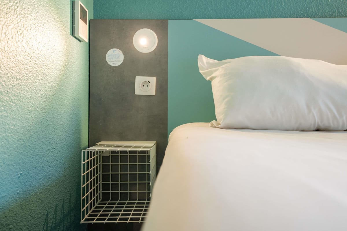 Une chambre d’hôtel bien rangée, avec un lit, une petite table de chevet, une lampe fixée au mur et une prise de courant.