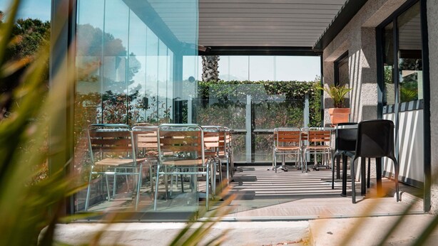 Un patio moderno al aire libre con paredes de vidrio, sillas de metal y una planta en maceta.