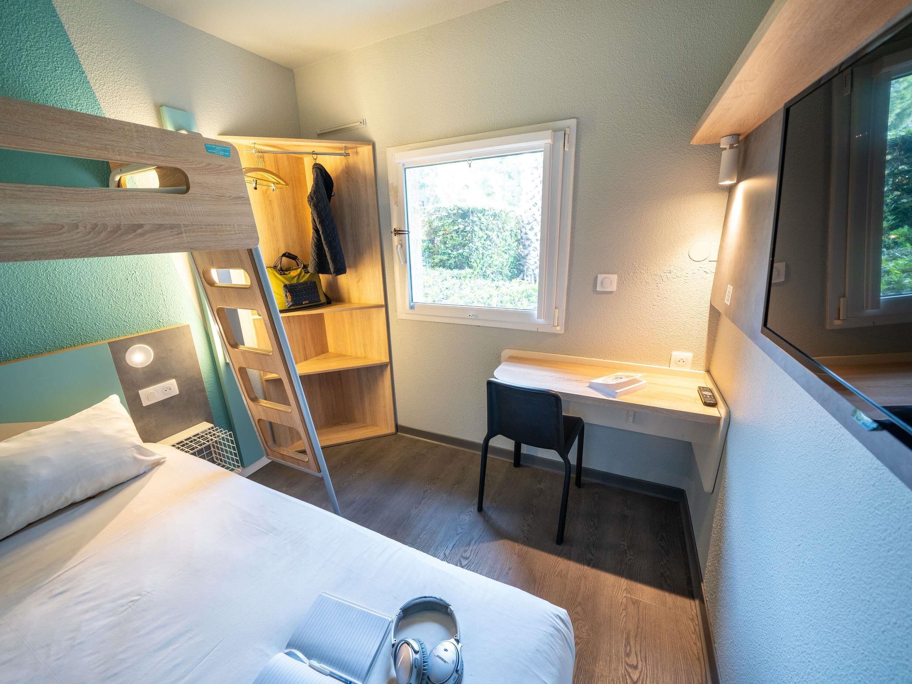 A compact room with bunk beds, a desk, a chair, a ladder, a window, and a wardrobe.