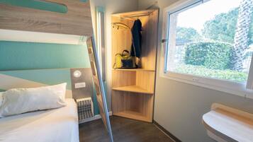 Een kleine kamer met een stapelbed, een klein houten plankje met een jas en een tas, een raam met uitzicht op groen, en een nachtkastje.