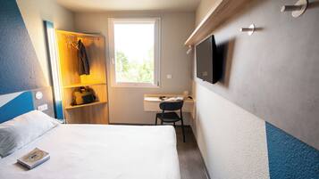 A hotel room with a bed, a desk, a chair, a TV mounted on the wall, and a window providing natural light.