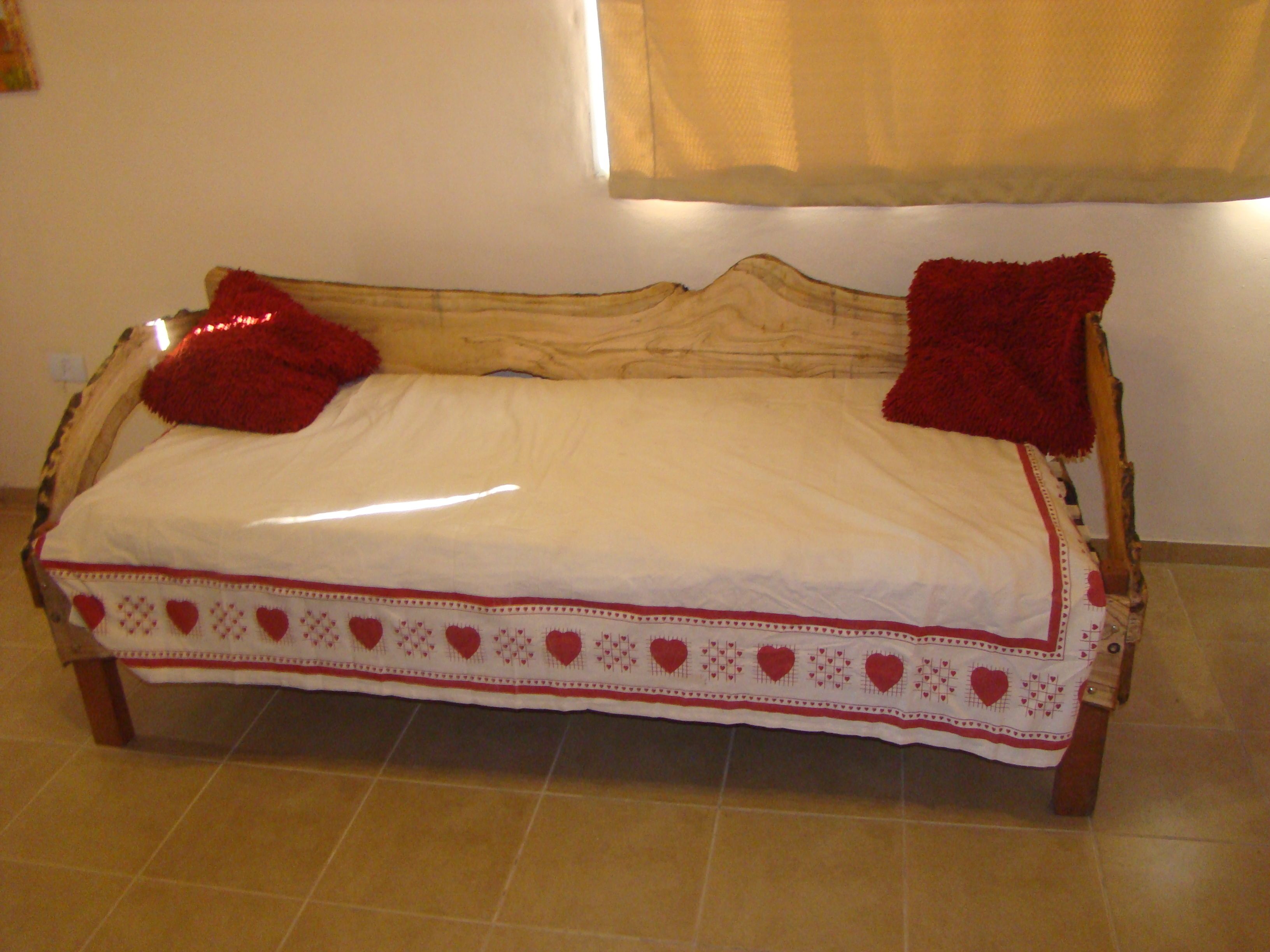 Ein Bett mit weißer Matratze und einer Bettdecke mit Herzmuster, einem Holzkopfende und zwei roten Kissen.