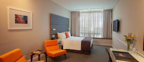 A hotel room with a bed, two orange chairs, a desk, a television, and a framed picture on the wall.