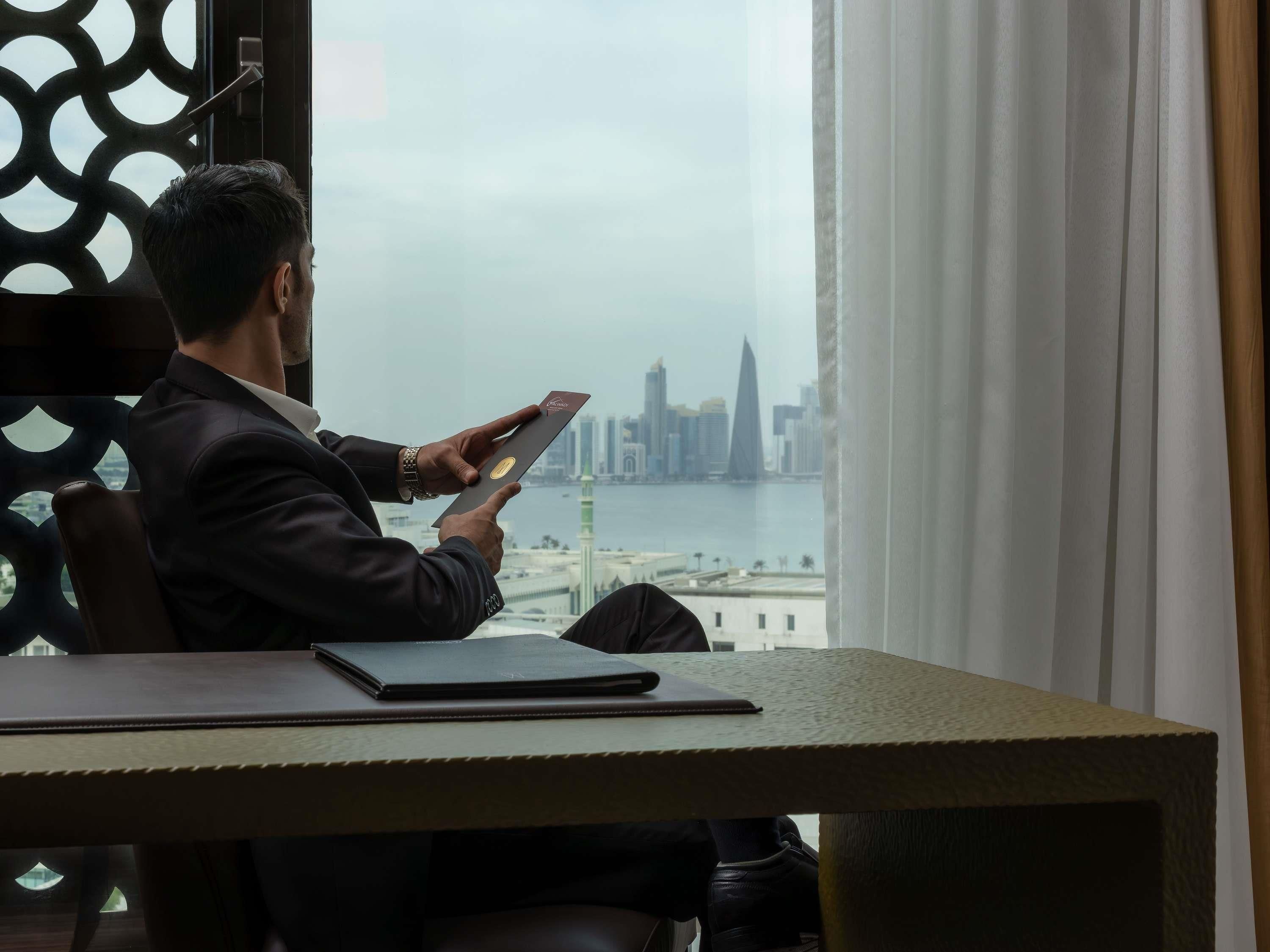 A man sitting at a table with a laptop, looking out at a cityscape with a river and buildings.