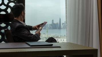 A man sitting at a table with a laptop, looking out at a cityscape with a river and buildings.
