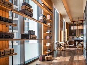 A modern hotel interior with wooden shelves displaying luggage, a dining area with chairs, and large windows.