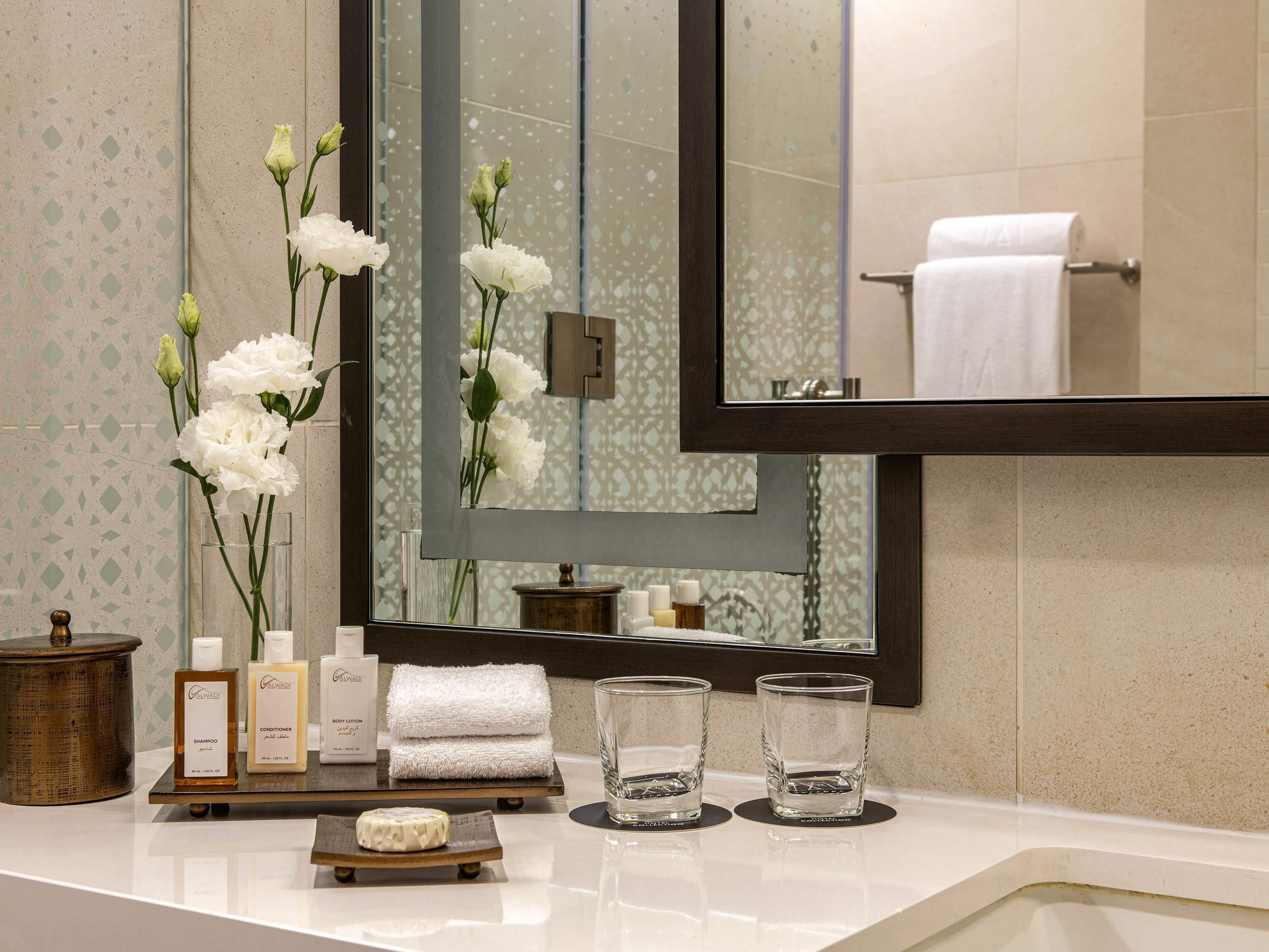 A bathroom vanity with a mirror, toiletries, and a vase of white flowers.