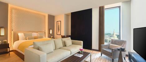Habitación de hotel con cama, sofá, mesa de centro, televisor y un ventanal con vista a la ciudad.