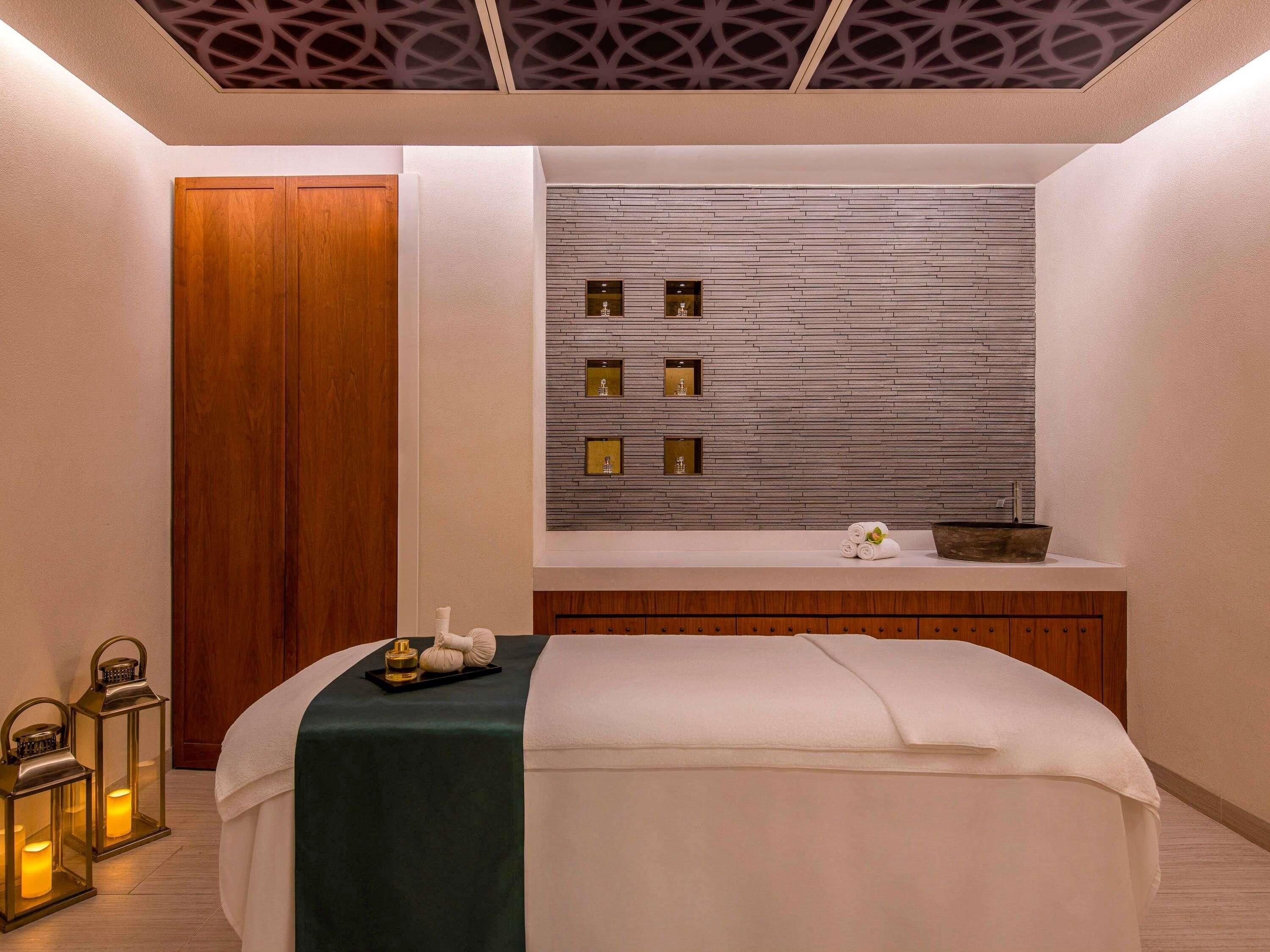 A spa room with a wooden door, a bed with a green towel, a wall with framed pictures, and a small shelf with a bowl and towels.