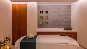A spa room with a wooden door, a bed with a green towel, a wall with framed pictures, and a small shelf with a bowl and towels.