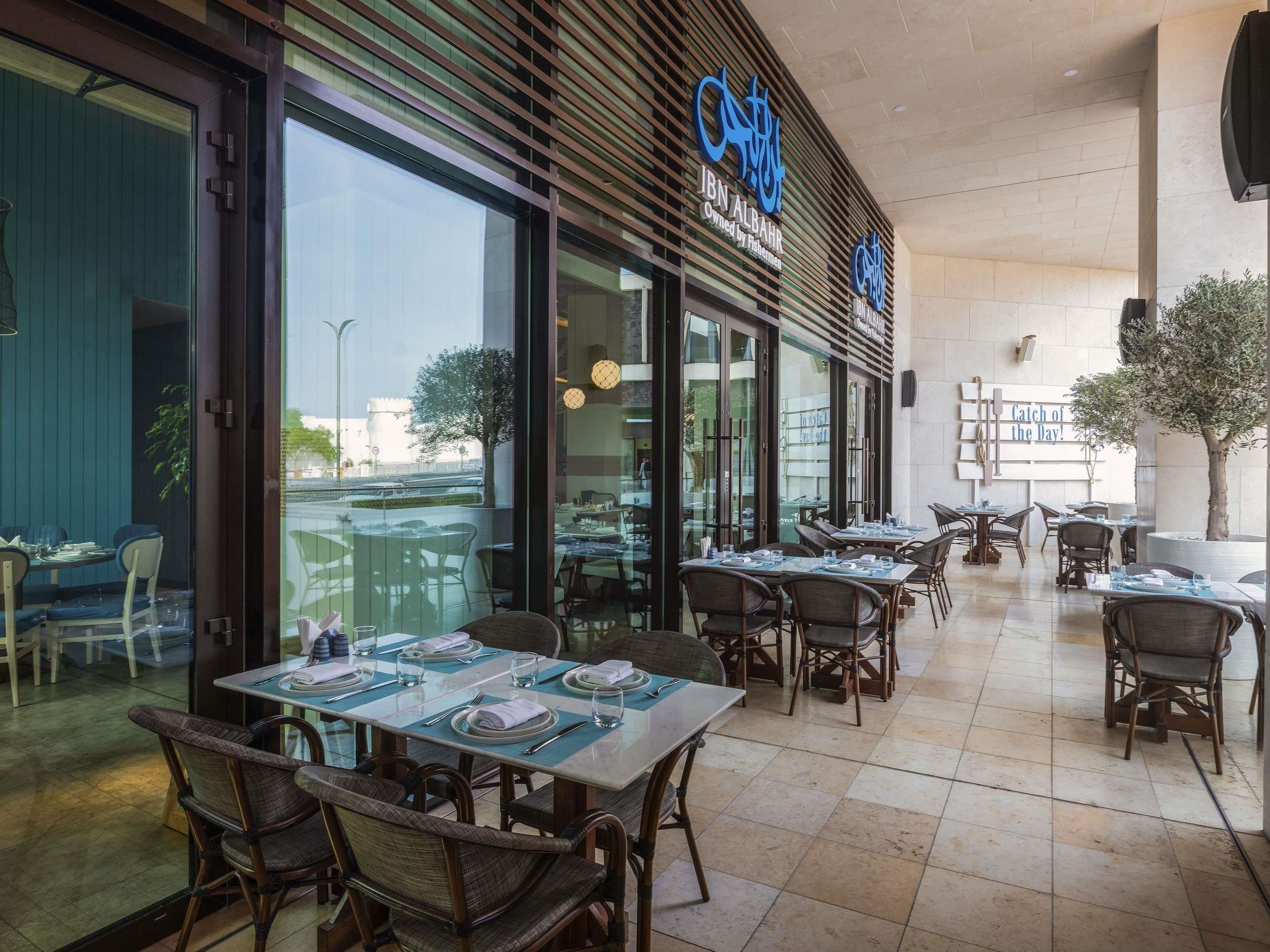 Un espace extérieur aménagé pour les repas, avec des tables et des chaises, une grande fenêtre et une enseigne indiquant « JBN ALBAHR ».