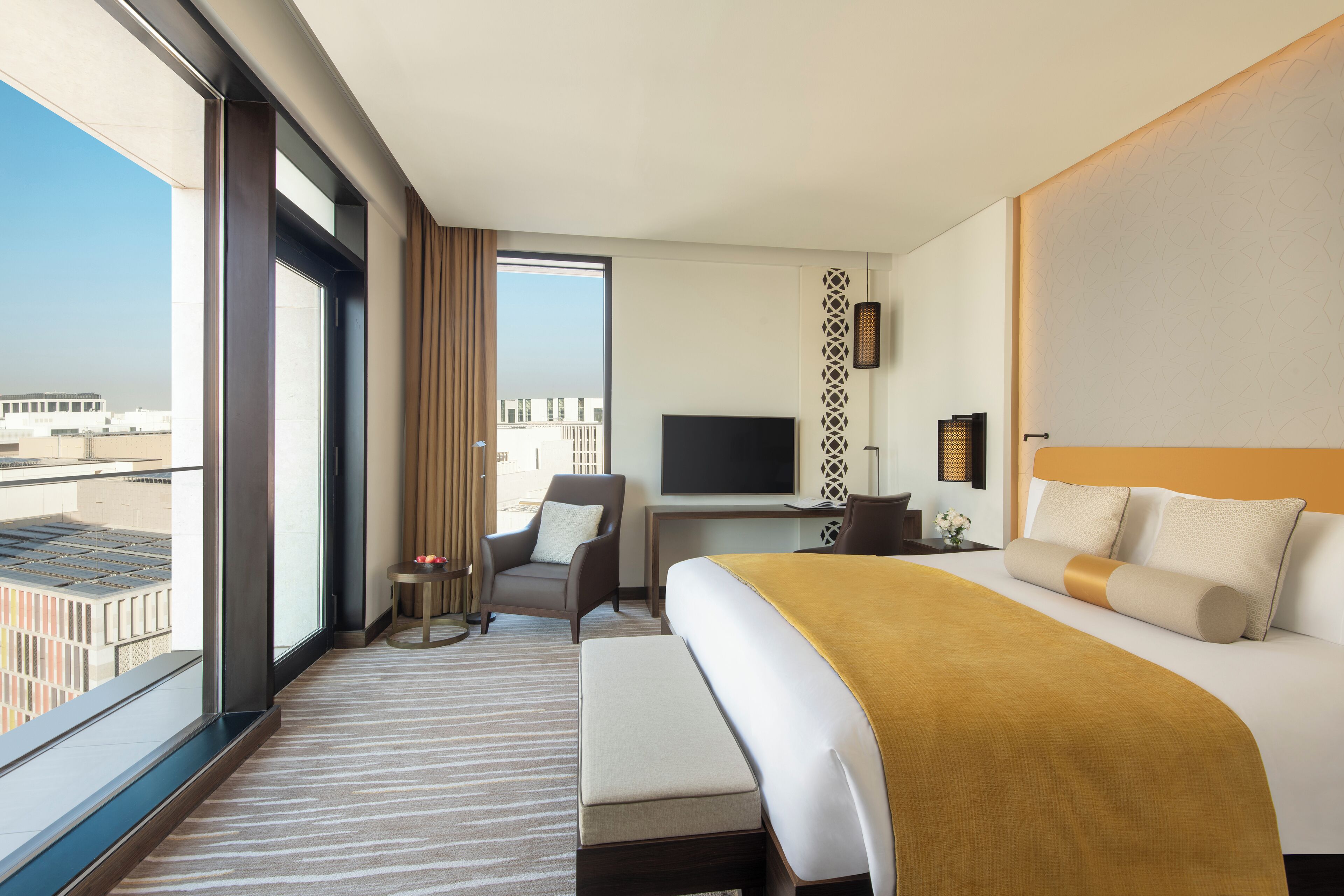 A hotel room with a large bed, a desk with a TV, a chair, and a view of the city through large windows.