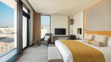 A hotel room with a large bed, a desk with a TV, a chair, and a view of the city through large windows.