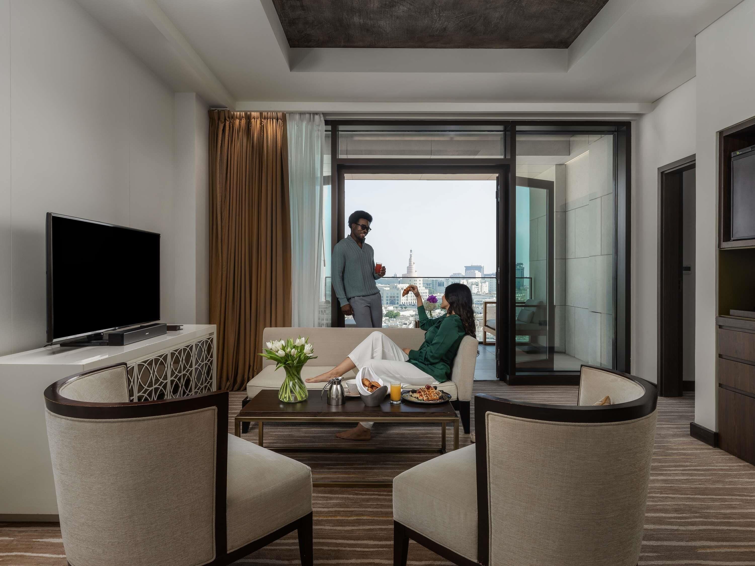 Een moderne hotelkamer met een flatscreen televisie, een bank, een bijzettafeltje met bloemen en een balkon met uitzicht op de stad.