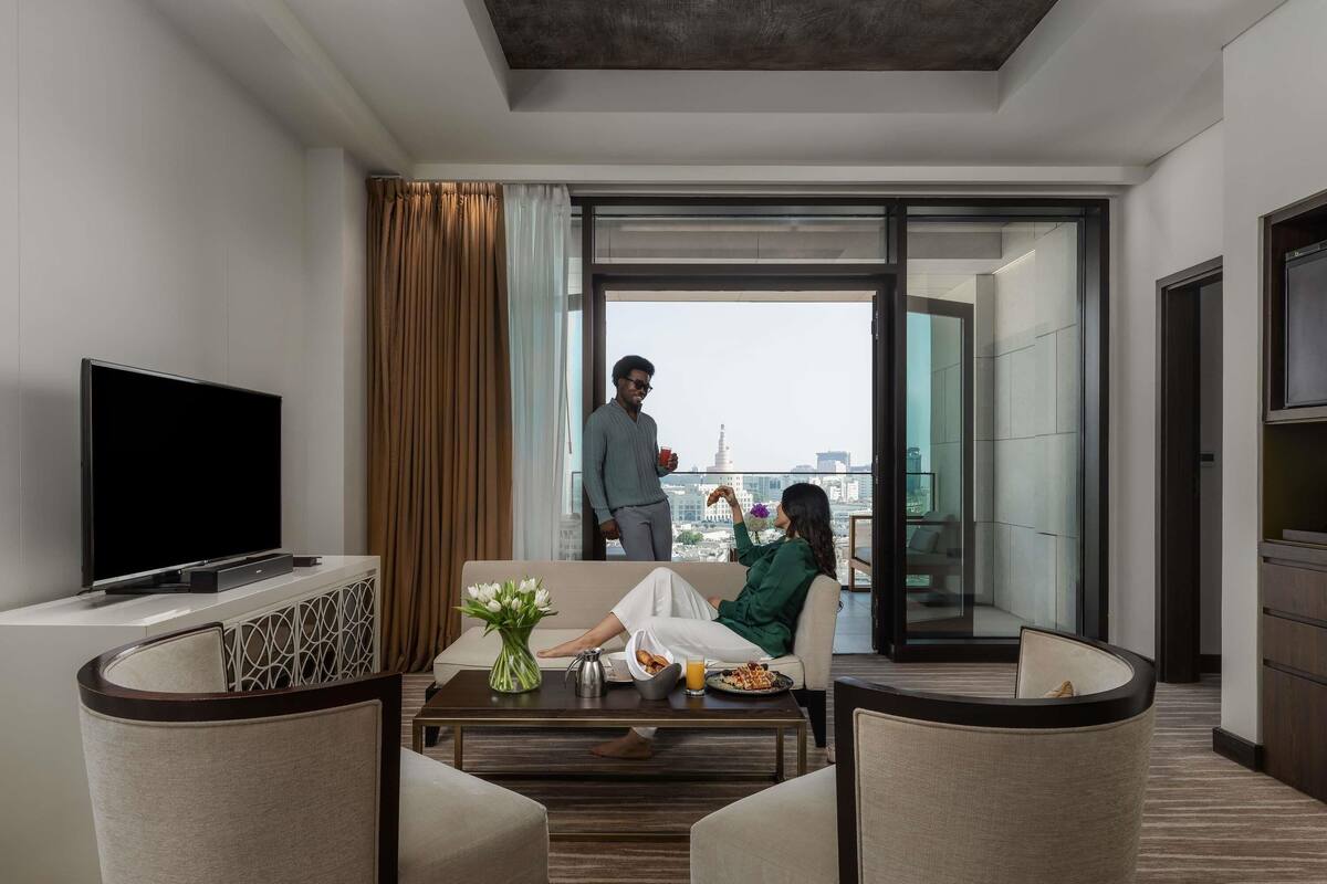 Une chambre dâhĂŽtel moderne dotĂ©e dâune tĂ©lĂ©vision Ă Ă©cran plat, dâun canapĂ©, dâune table basse agrĂ©mentĂ©e de fleurs et dâun balcon offrant une vue sur la ville.