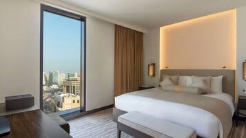 Une chambre d’hôtel avec un grand lit, un bureau, une chaise, une table de chevet, une lampe fixée au mur et une vue sur la ville par une grande fenêtre.