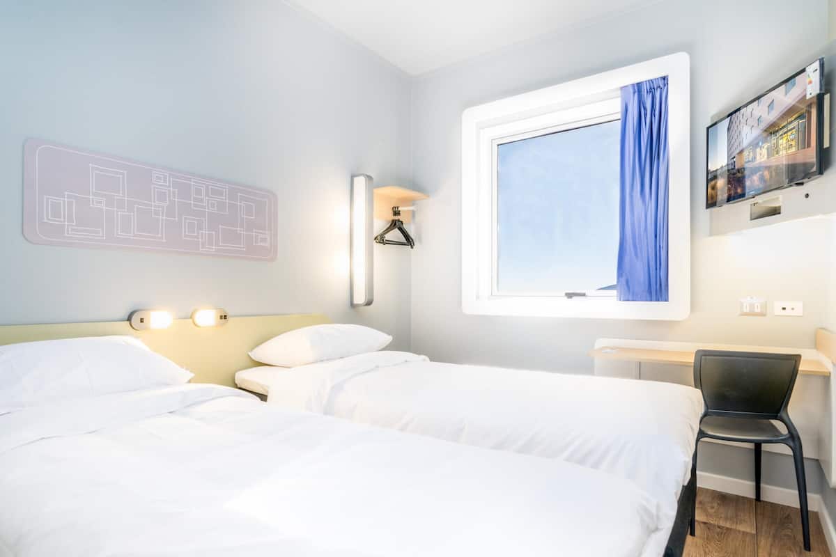 A hotel room with two beds, a desk, a chair, a window with a curtain, wall-mounted lights, and a wall art piece.