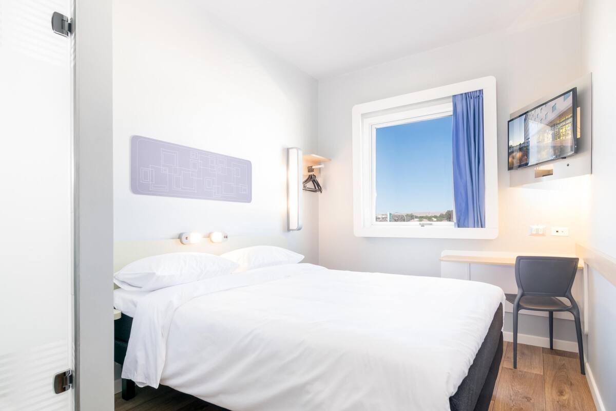 A bedroom with a bed, a desk, a chair, a window with a view, and a wall-mounted light fixture.