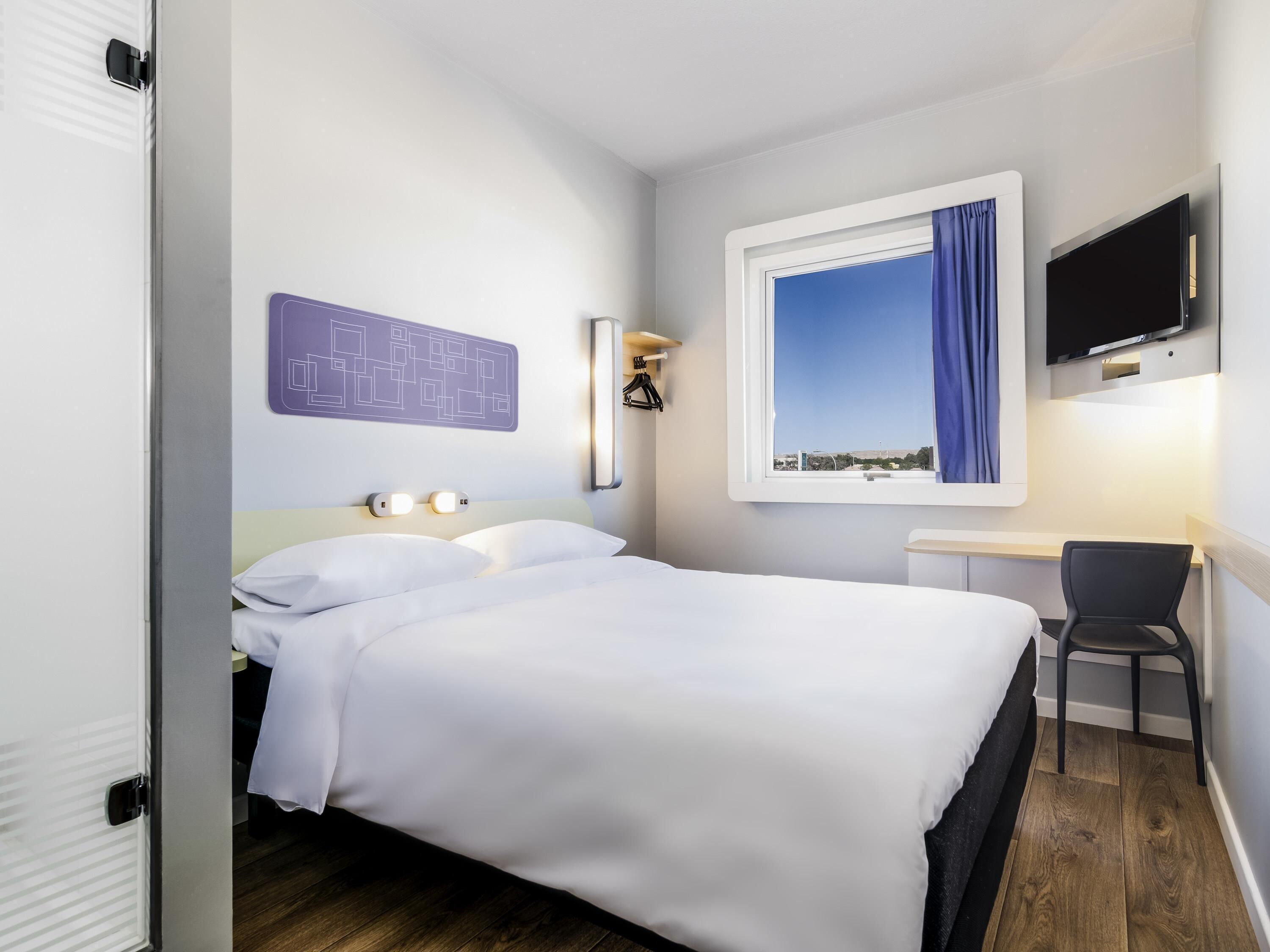 Habitación de hotel con cama, escritorio, silla, televisor montado en la pared y ventana con cortina.