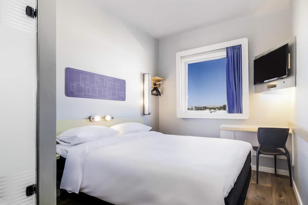 Habitación de hotel con cama, escritorio, silla, televisor montado en la pared y ventana con cortina.