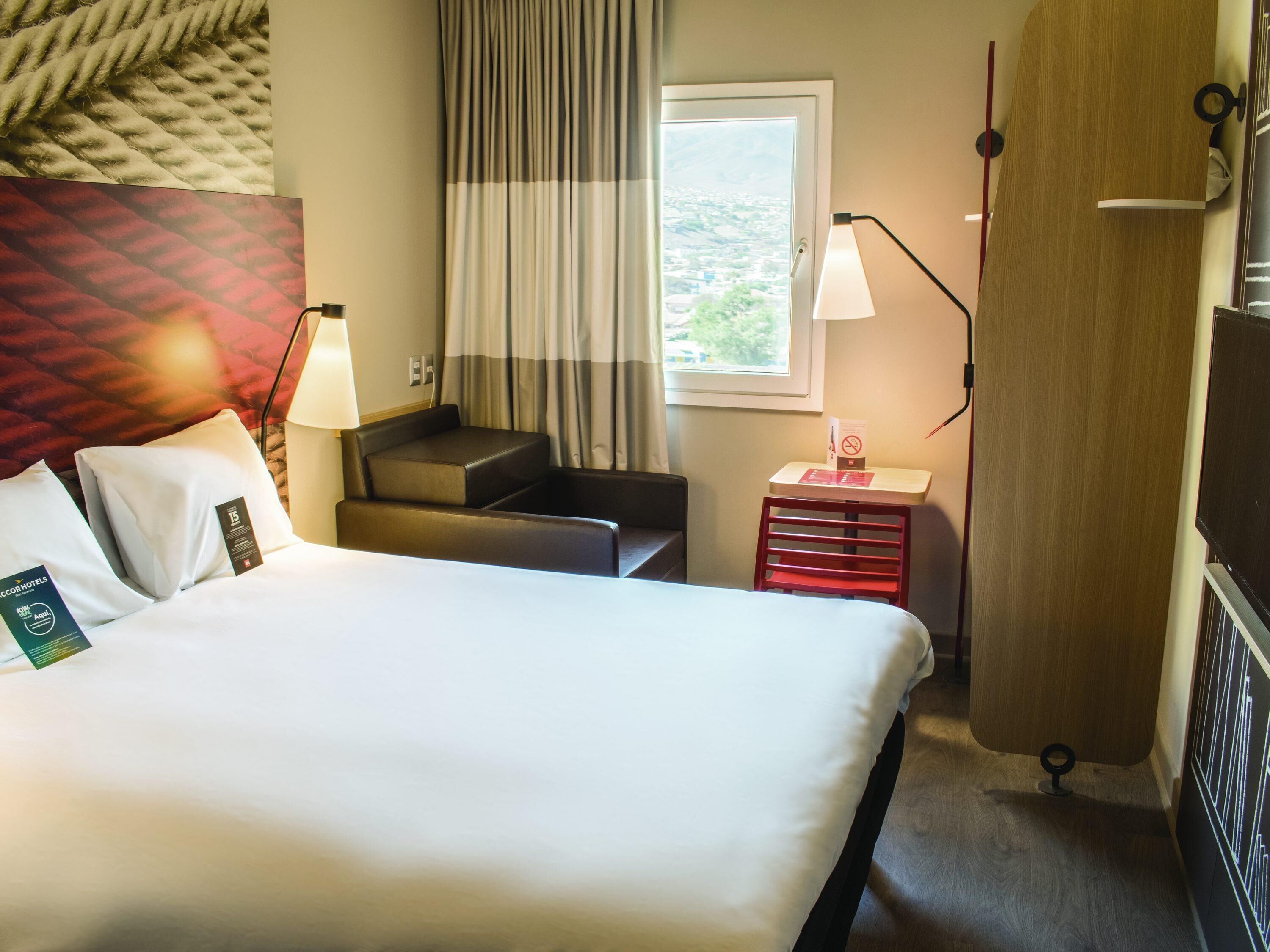 Habitación de hotel con cama, escritorio, silla, ventana con cortinas y lámpara.