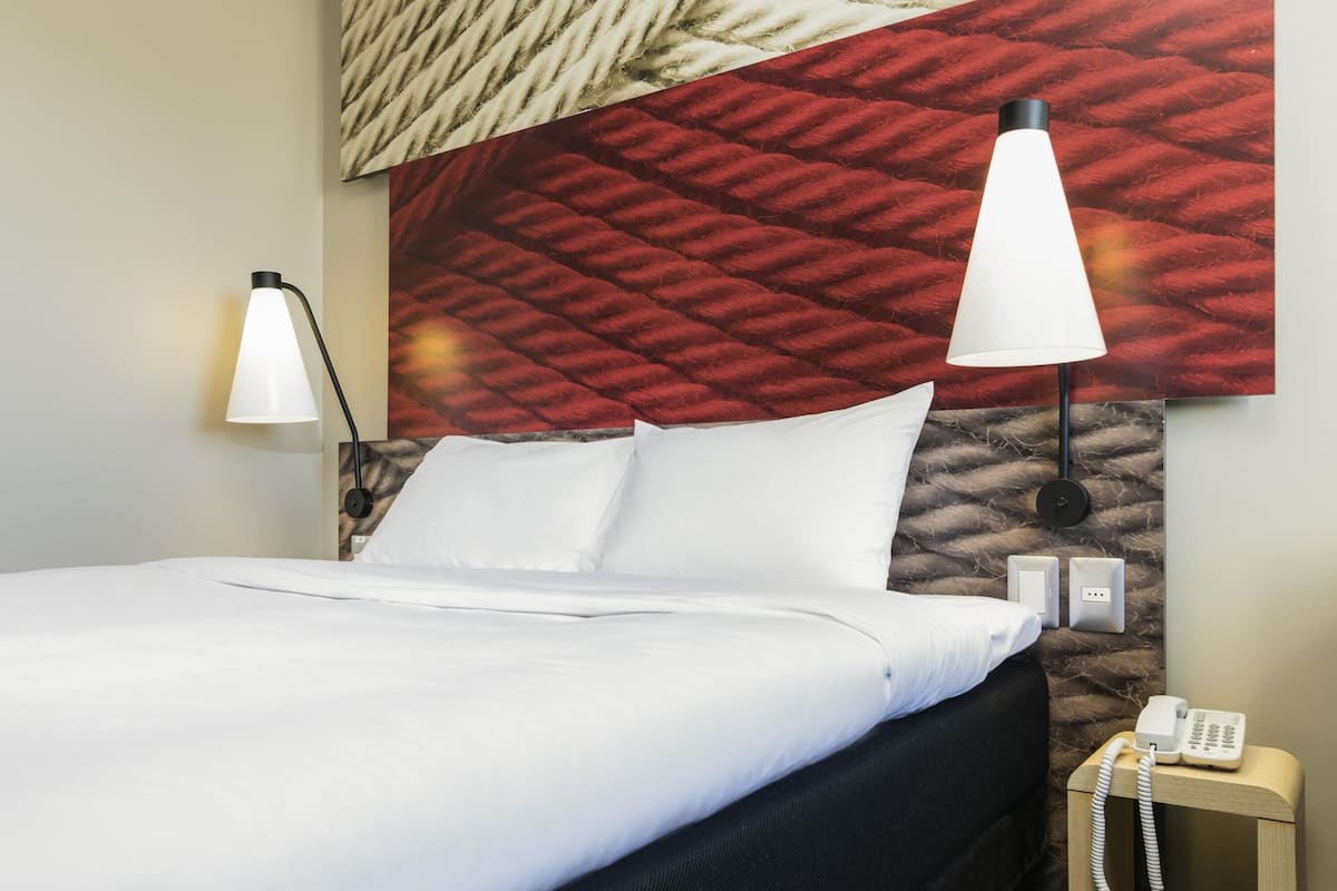 Una habitación de hotel con una cama, dos lámparas de noche, un teléfono y una pared texturizada detrás de la cama.