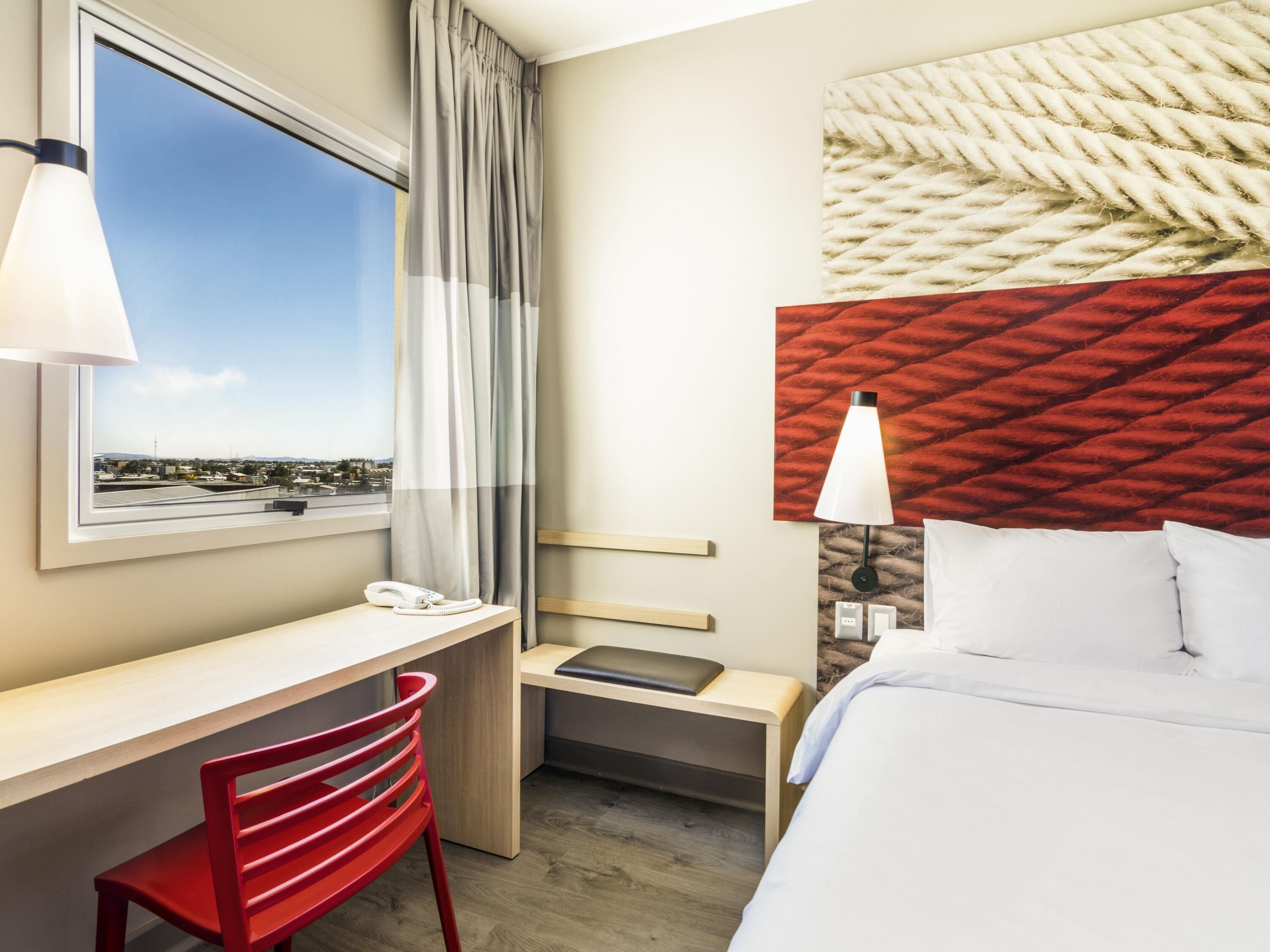 Habitación de hotel con escritorio, silla, cama y vista del paisaje urbano a través del ventanal.