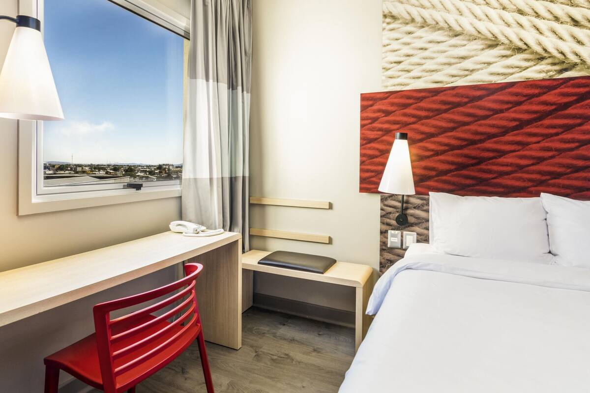 Habitación de hotel con escritorio, silla, cama y vista del paisaje urbano a través del ventanal.