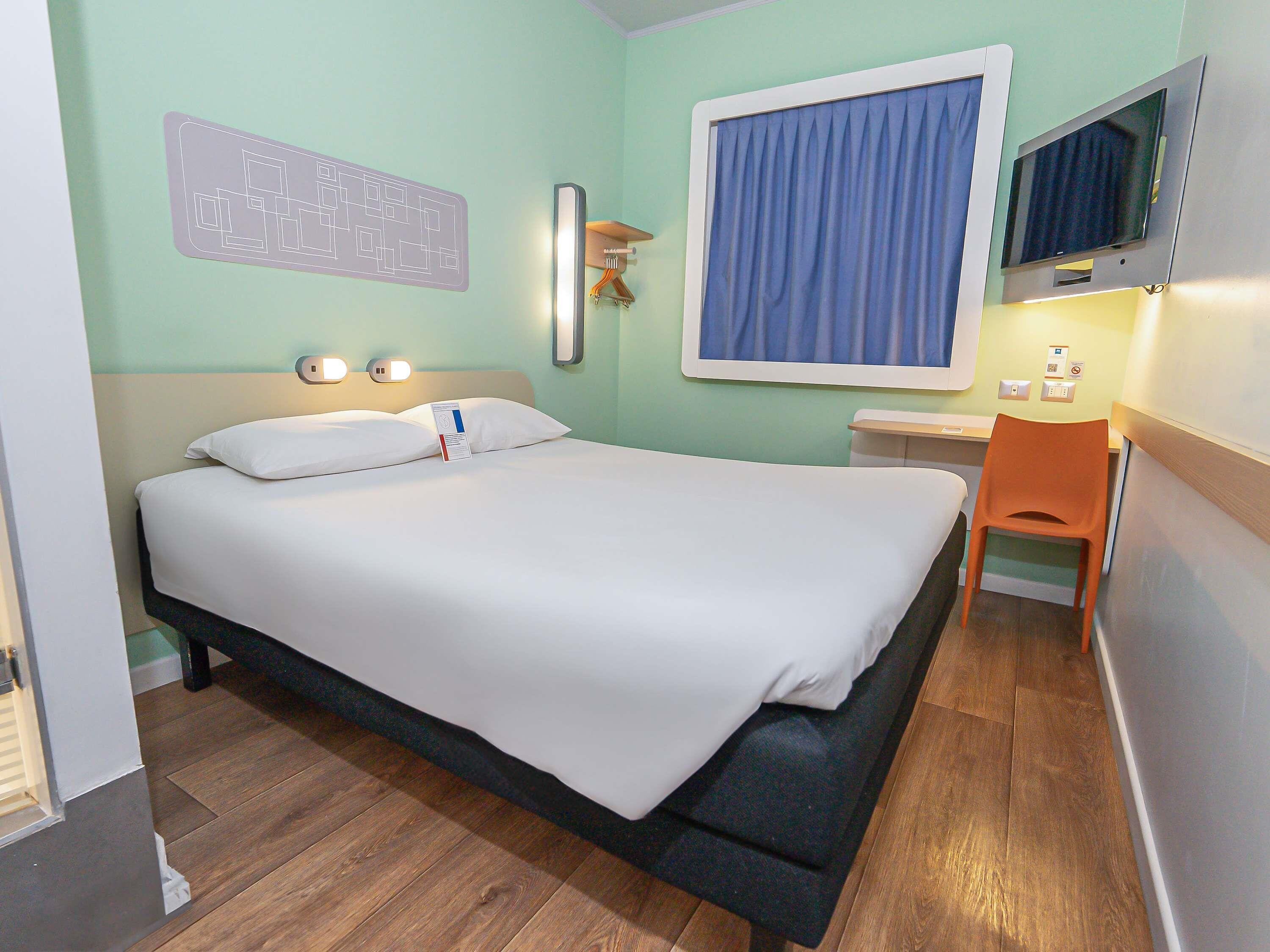 A hotel room with a bed, a desk, a chair, a television, and a wall-mounted light fixture.